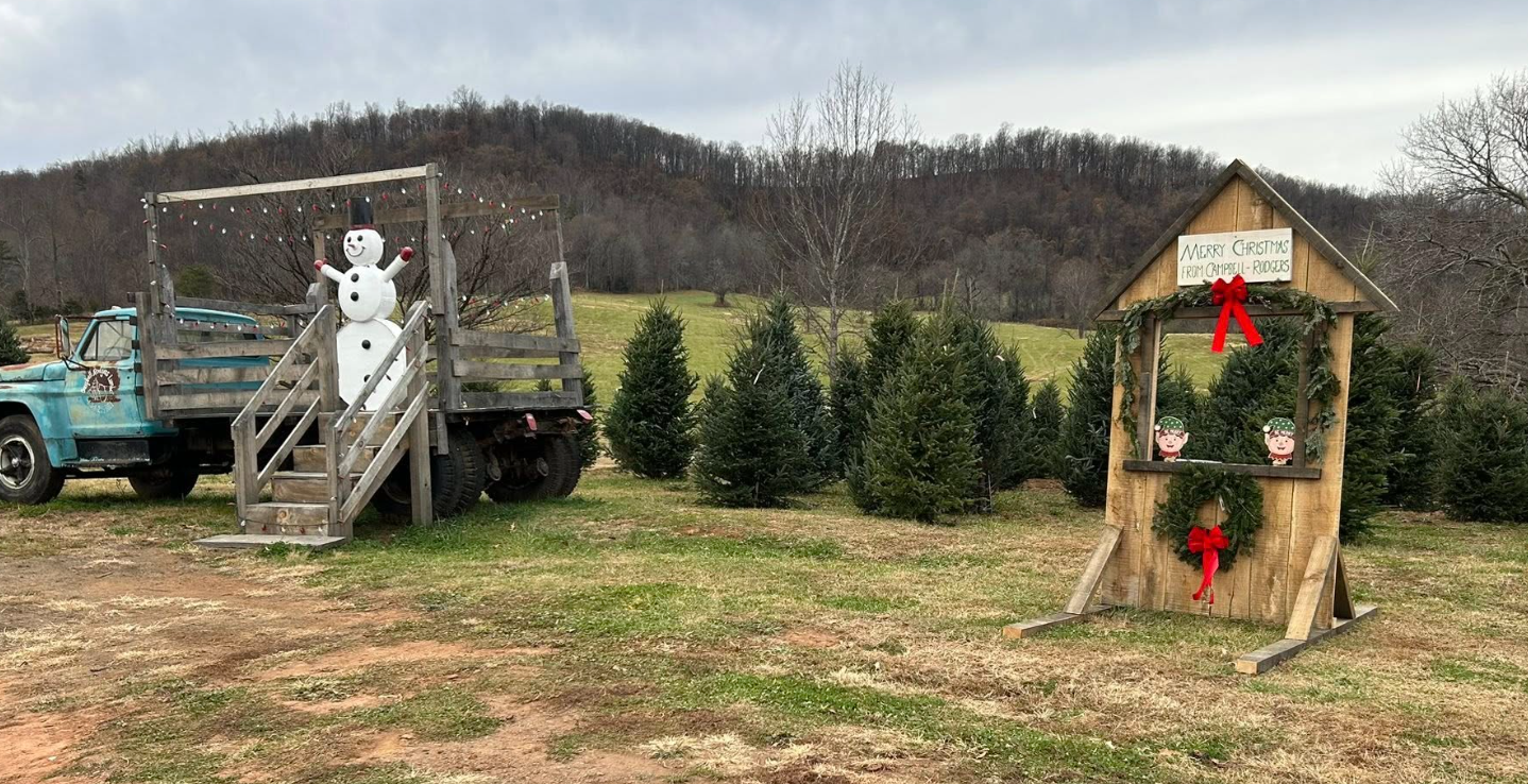 A decorated Christmas scene outdoors with a vintage truck and a snowman on a wooden platform, and a festive board with Christmas decorations and a sign saying 'Merry Christmas from Campbell-Rodgers' amidst Christmas trees.