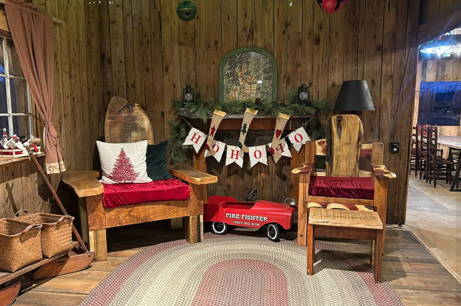 Cozy indoor holiday scene with wooden furniture, a fireplace decorated with greenery and stockings, a 'HO HO HO' banner, and festive decorations including a throw pillow with a Christmas tree design, a red velvet seat cushion, and a toy fire truck.