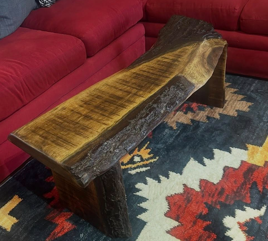 A wooden bench made from a thick, natural wood slab with a live edge, placed on a colorful patterned rug in front of a red upholstered sofa.
