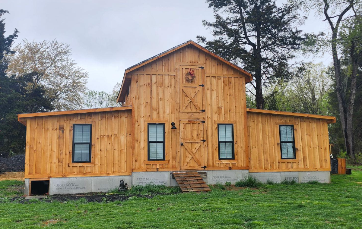 A wooden barn-style house under construction, with a concrete foundation, three windows with black frames, and a central barn door with a small step in front. The house is surrounded by grass and trees.
