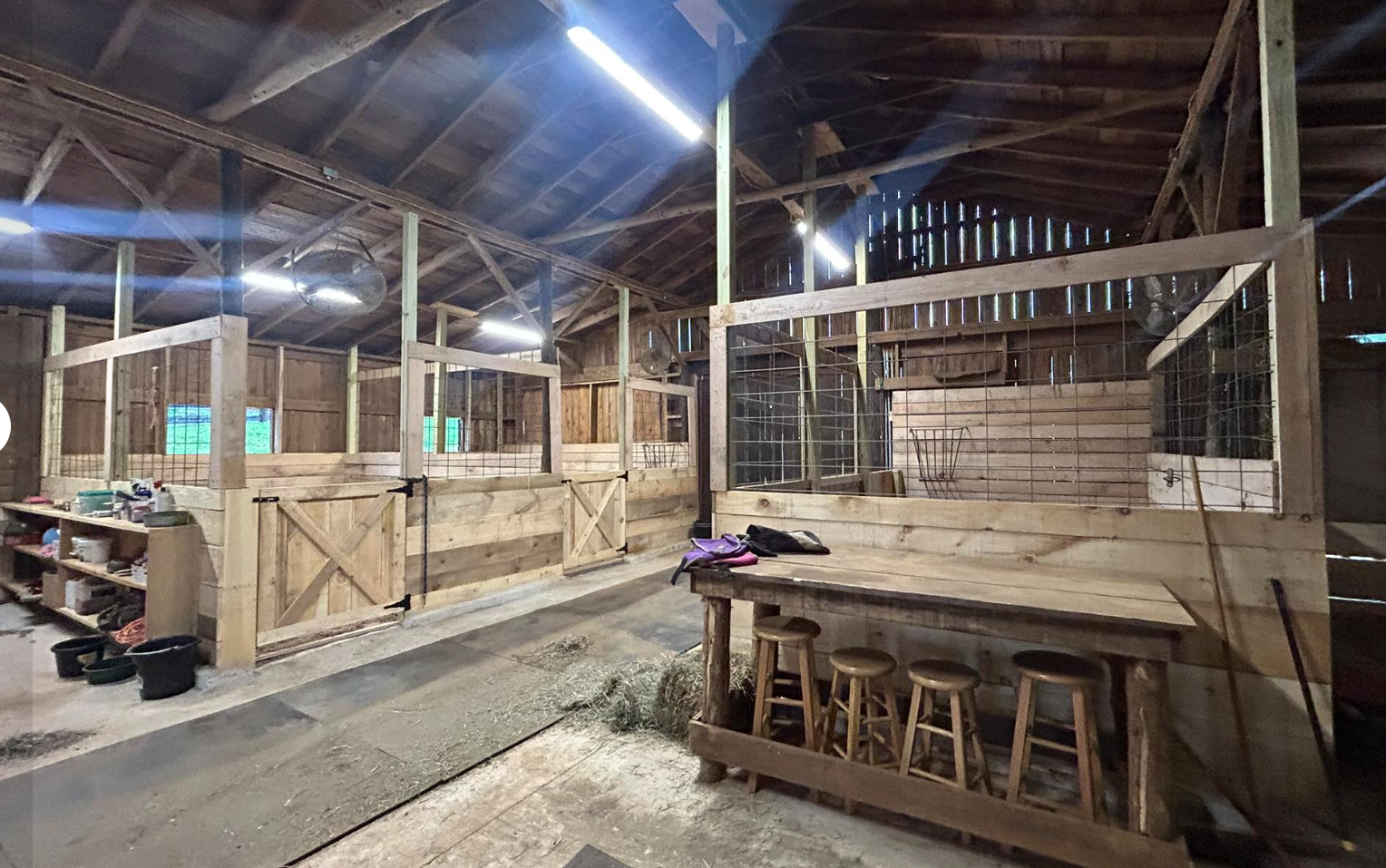 Interior of a barn with wooden stalls, some with gate doors, and a workspace with stools and a table. The barn has a high, open ceiling with exposed wooden beams and some light coming through gaps in the walls.