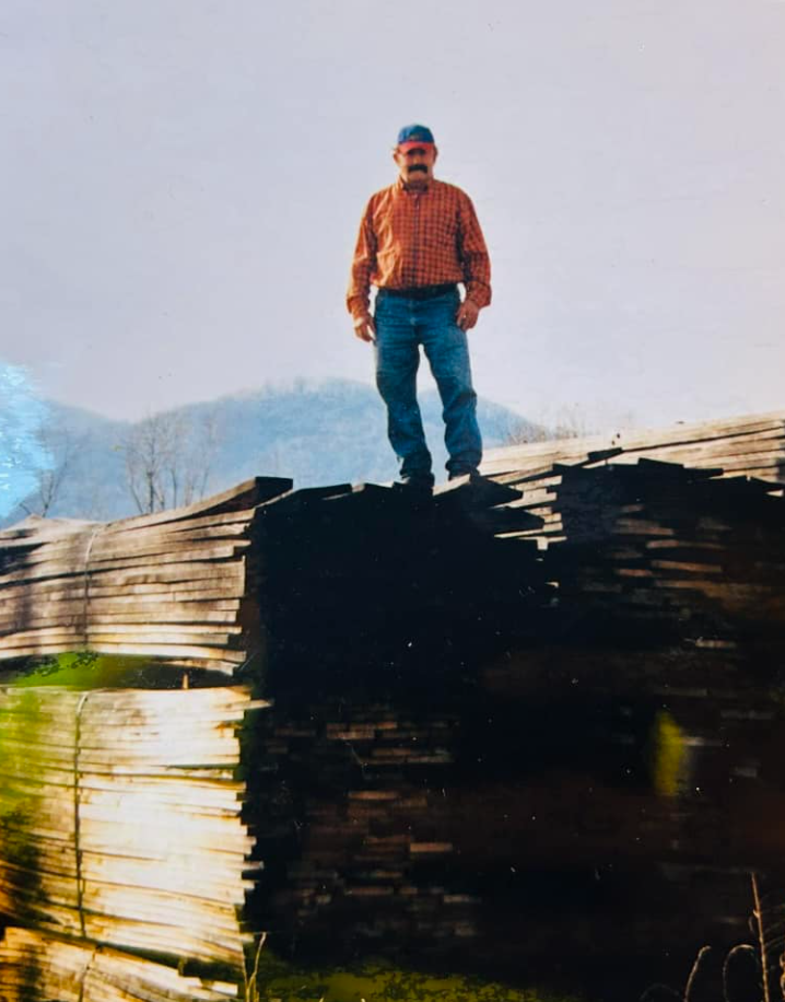 A man with a mustache dressed in a red plaid shirt, blue jeans, and a backwards blue cap stands on a pile of cut logs outdoors with hills and trees in the background.