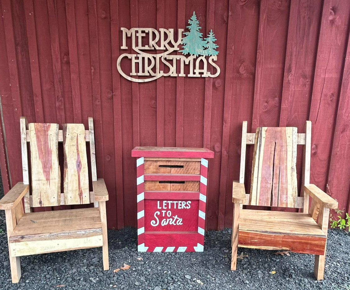 Two wooden chairs and a red mailbox labeled "Letters to Santa" are placed in front of a red wooden wall. A "Merry Christmas" sign with trees is hanging above them.