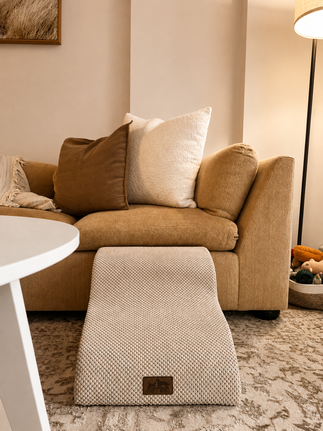 A living room with a beige sofa, decorated with two large pillows, a throw blanket, and a textured footrest labeled "Cozy Kiss." In the background, a floor lamp and a basket with toys are visible, with a framed picture hanging on the wall.