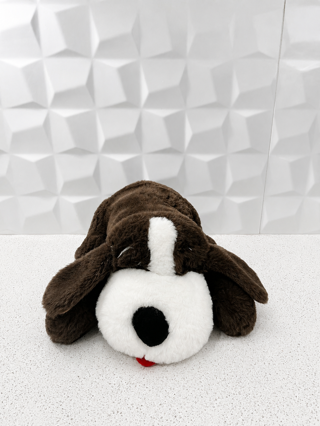 A plush stuffed dog toy with a white face, black nose, and long floppy ears, lying on a white surface against a geometric white tiled wall.