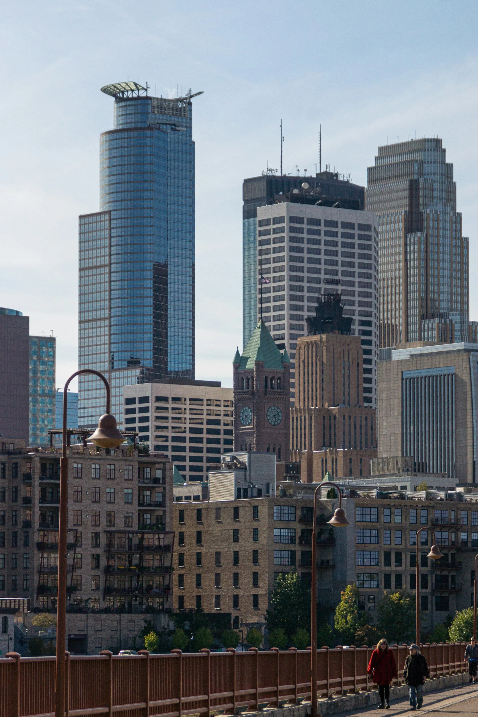 City skyline with modern skyscrapers and historic clock tower, people walking along a sidewalk with street lamps in the foreground.