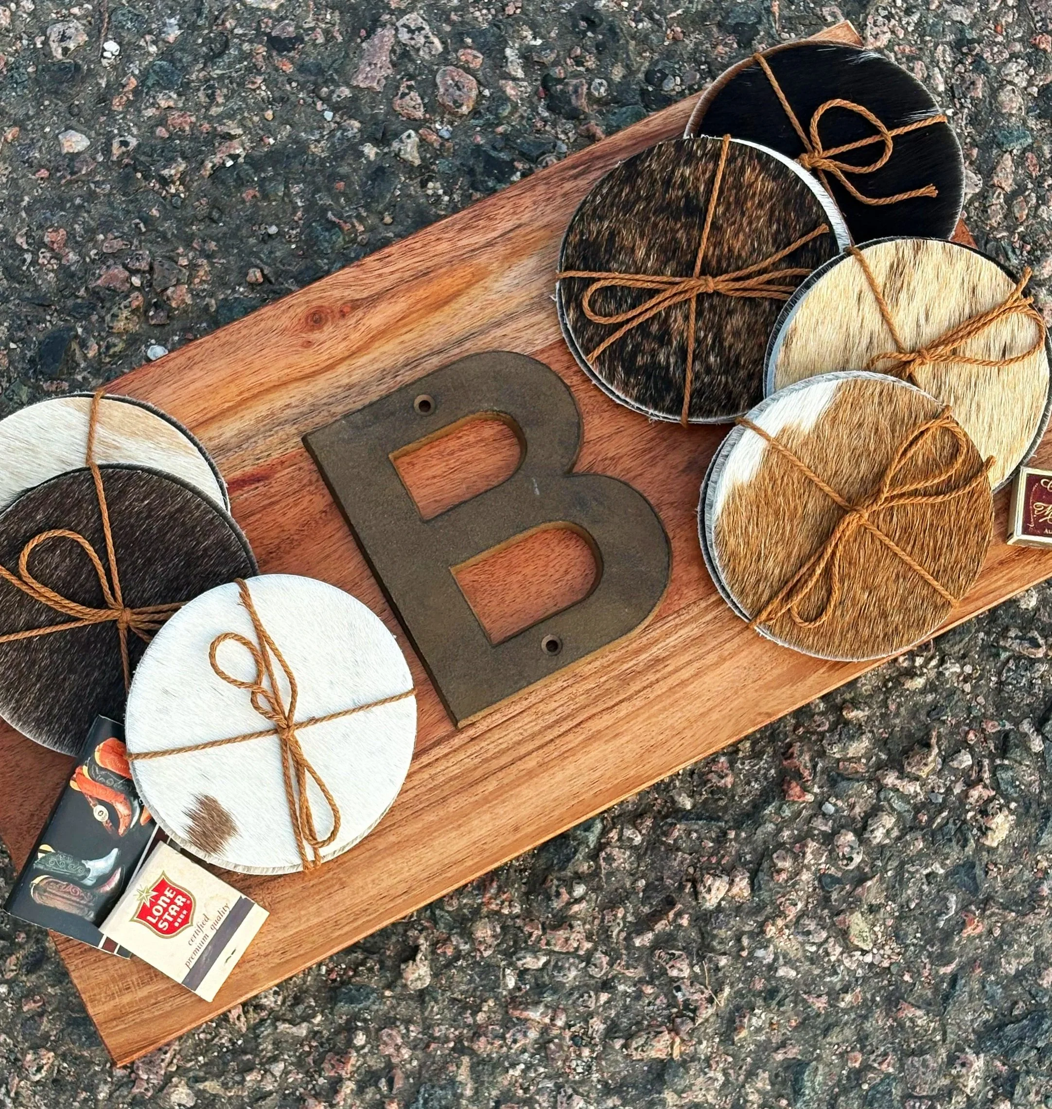 Several circular cowhide coasters tied with twine on a wooden board, with a metal letter B and a matchbook nearby, on a textured dark granite surface.