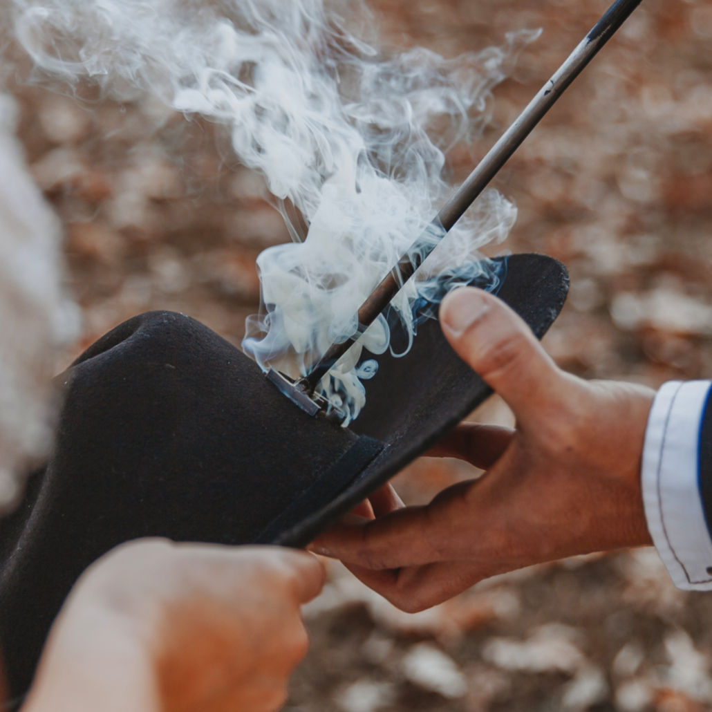 Close-up of a person holding a black cowboy hat branding initials with a smoking branding iron.