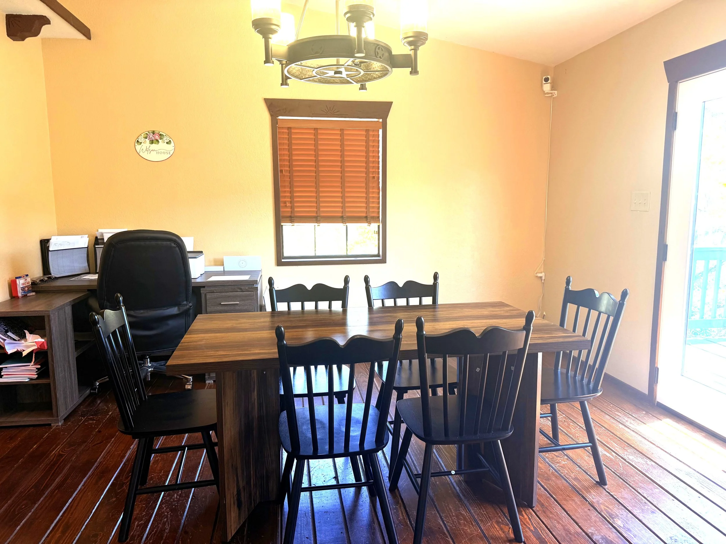 A dining room with a wooden table surrounded by black chairs, a window with wooden blinds, a corner desk with office supplies, a yellow wall with a small decorative sign, and a ceiling chandelier with multiple bulbs.