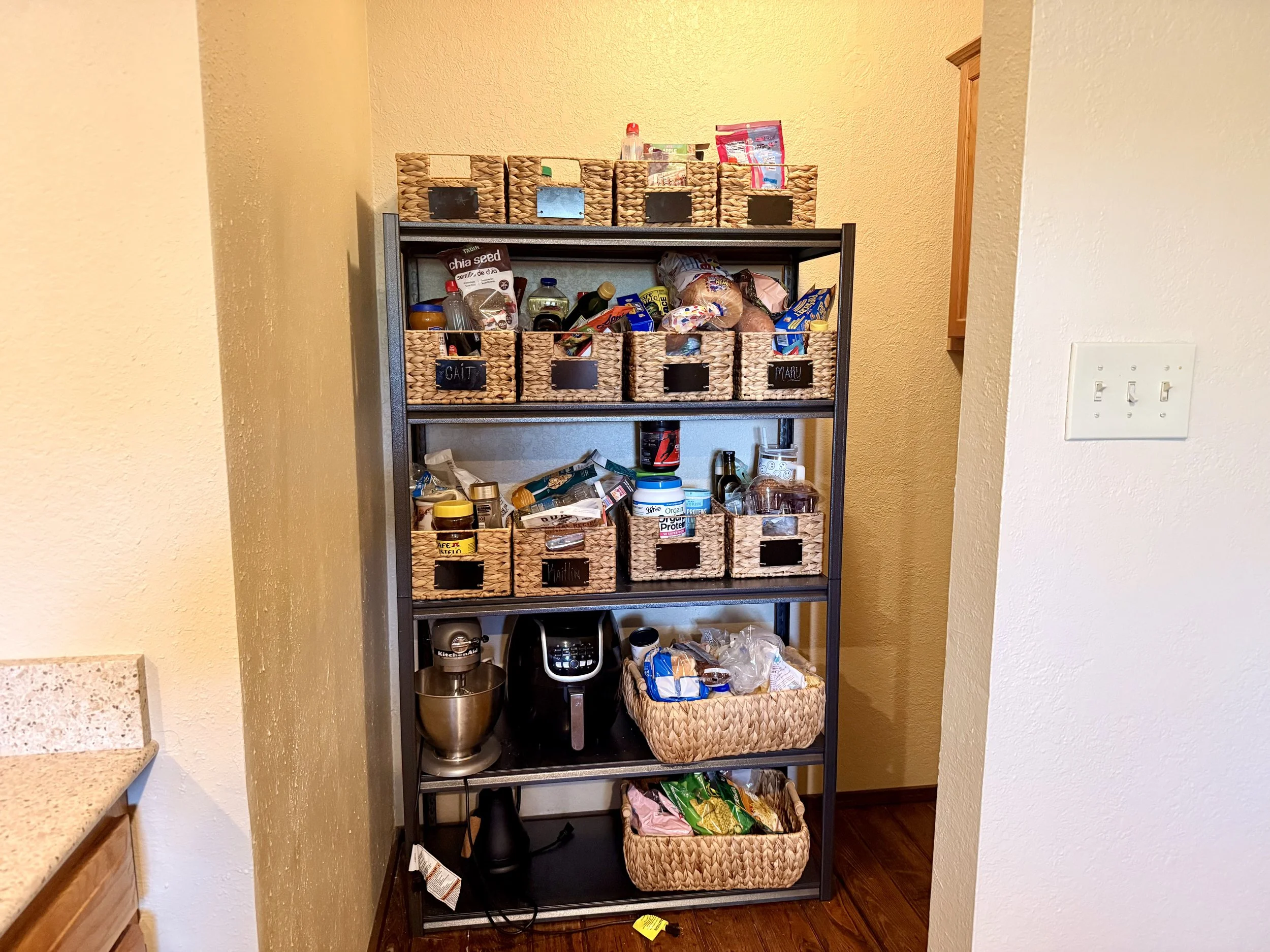 A black metal shelf with woven baskets containing food items, including snacks and spices, in a kitchen corner with beige textured walls and a light switch on the right.