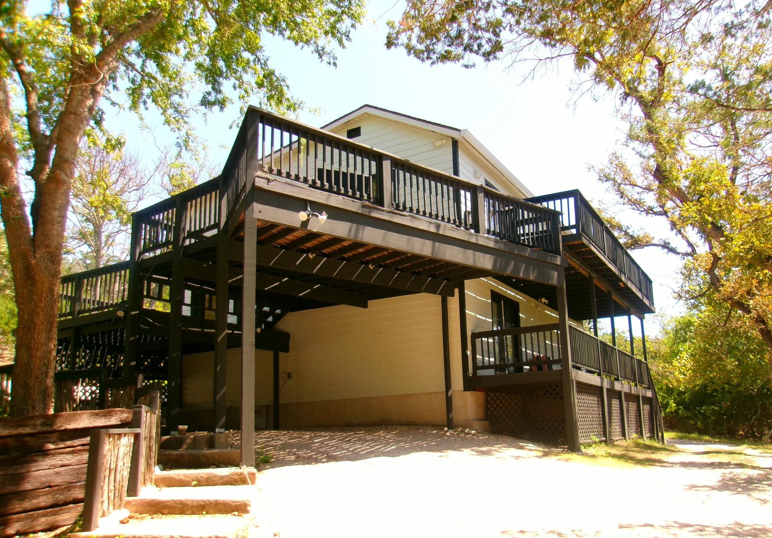 The image shows the underside of a wooden deck attached to a house, surrounded by trees with green leaves, and a clear sky overhead.