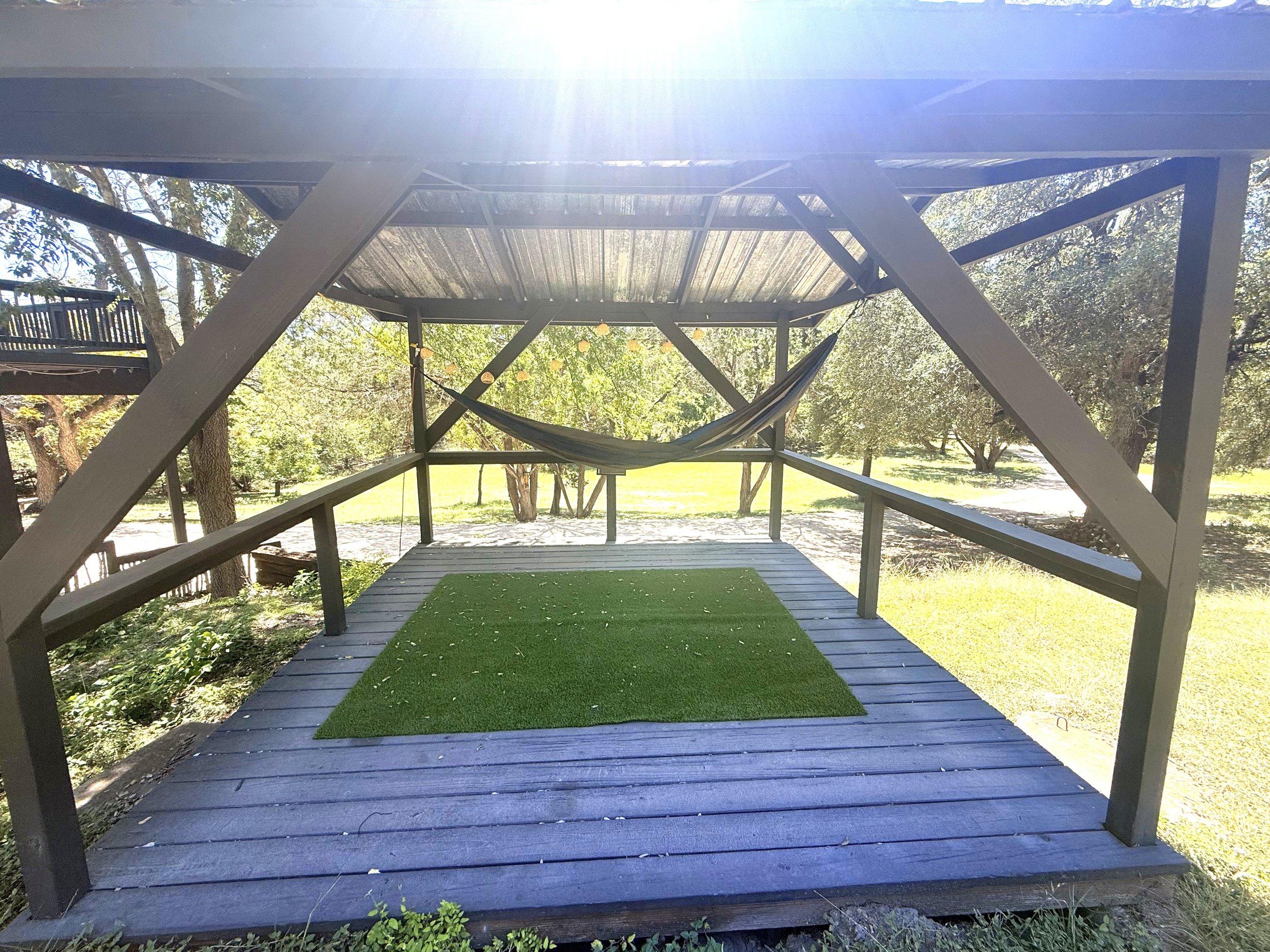 A raised wooden deck with a black hammock hanging underneath. There is a green artificial grass mat on part of the deck. The deck is in a park or backyard with trees and grass surrounding it. The roof of the deck is made of wood and is open at the si