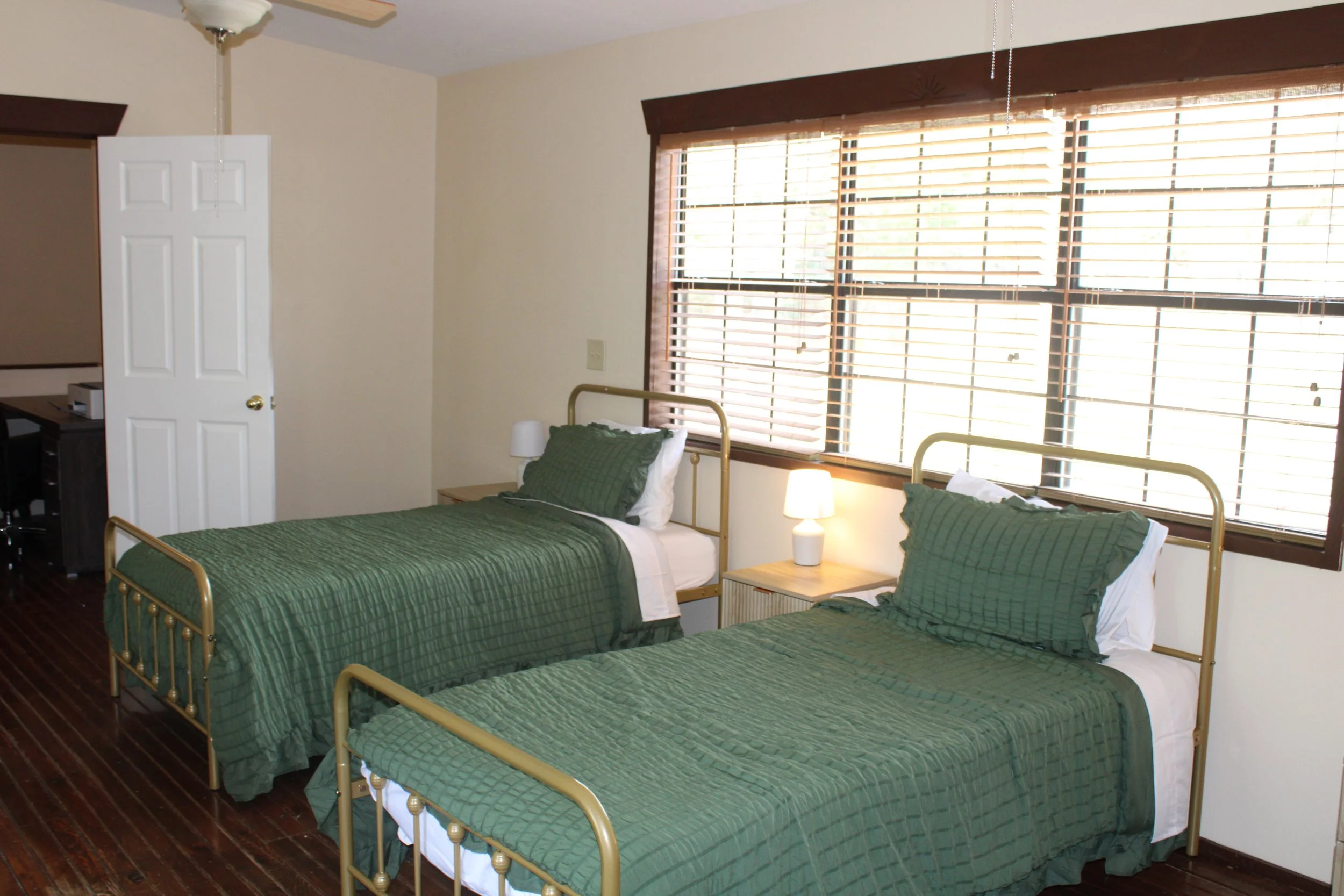 A bedroom with three beds covered in green quilts, pillows, and white sheets. Each bed has a small nightstand with a white lamp. We provide bedding