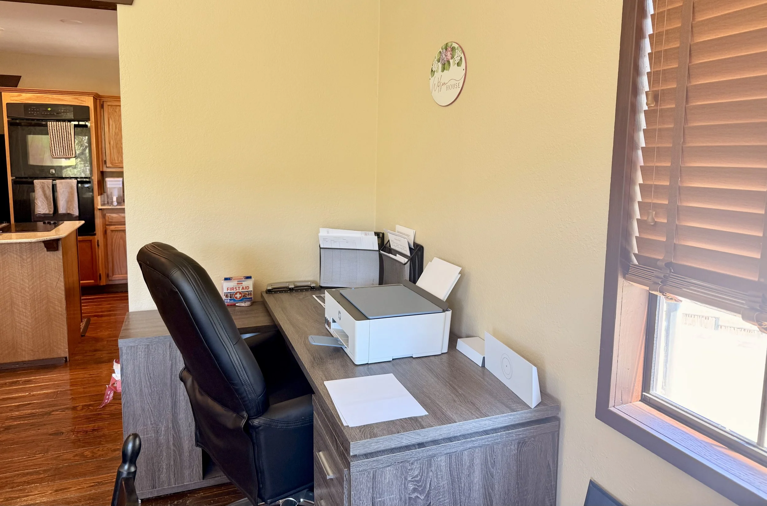 A home office with a desk, a black office chair, and various office supplies including a printer, papers, and folders. There is a window with blinds and a yellow wall with a decorative sign that reads 'Vigufur House'.