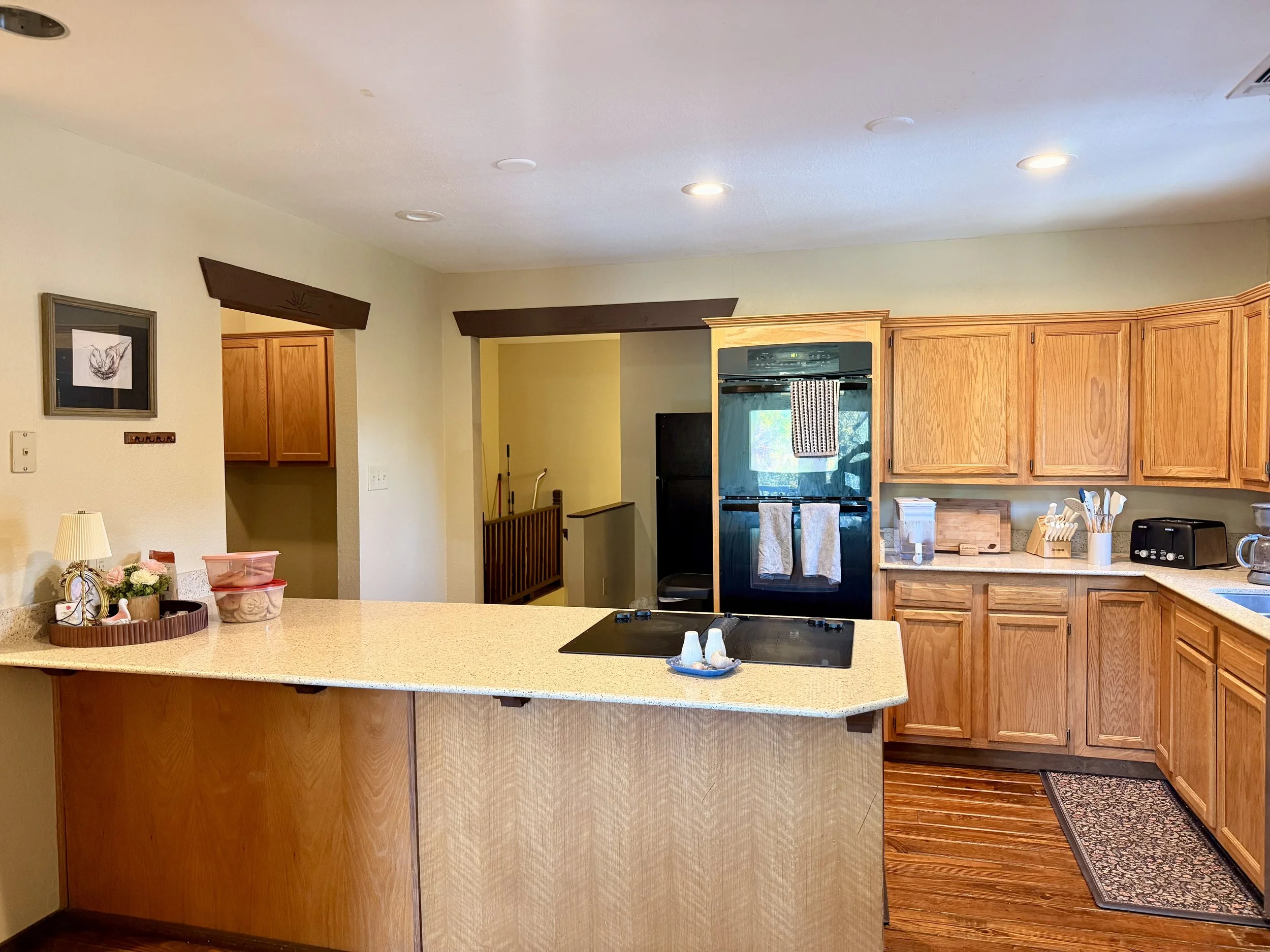 A kitchen with wooden cabinets, a black oven, and a beige speckled countertop. Items on the counter include a small lamp, flowers, a pink container, a cutting board, and a toaster. Recessed ceiling lights and a window with a towel hanging are visible.
