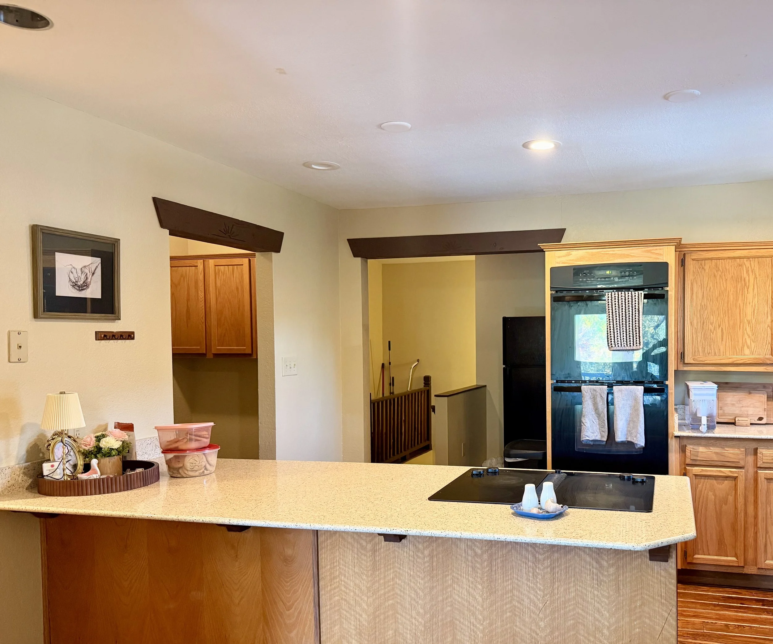 Kitchen with beige countertop, wooden cabinets, black stove, and a yellow wall. Decor includes a lamp, a picture frame, a floral arrangement, and containers on the counter. Ceiling with recessed lighting.