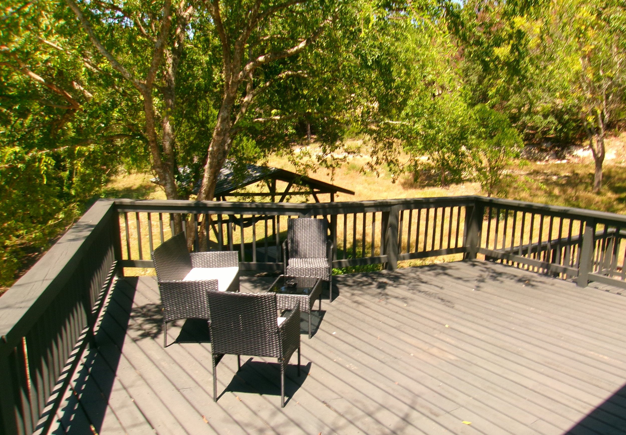A wooden deck with black patio furniture including a table and several chairs, surrounded by green trees and sunlight filtering through the leaves.