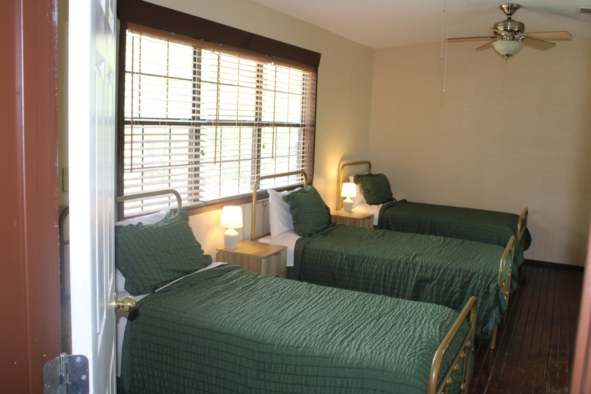 A bedroom with twin beds covered in green quilts, pillows, and white sheets. Each bed has a small nightstand with a white lamp. We provide bedding