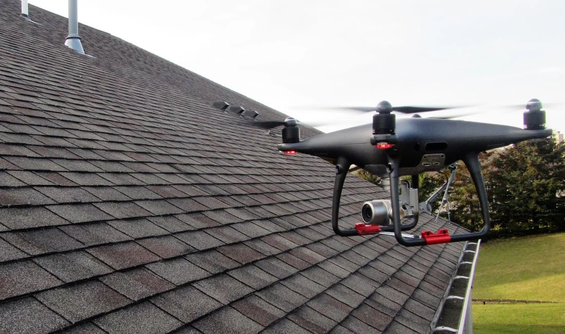 A drone flying on a house roof with gray shingles, with a camera mounted underneath.
