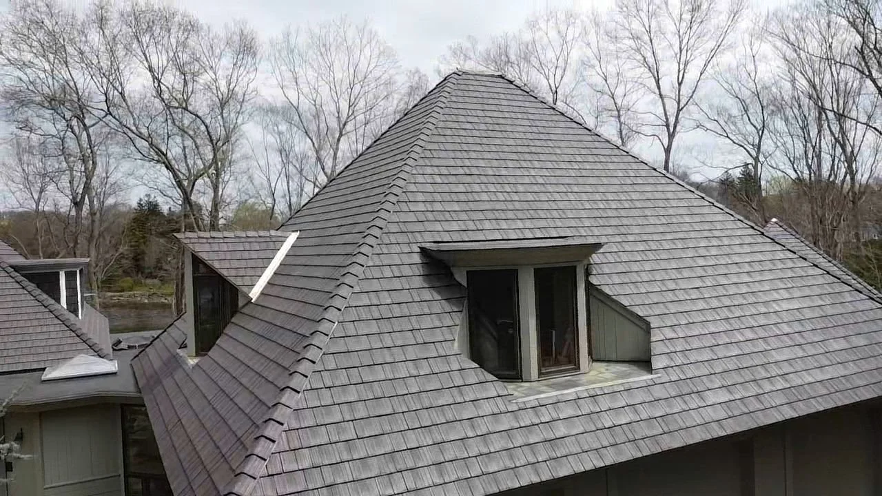 A house rooftop with gray shingles and a central dormer window, surrounded by leafless trees.