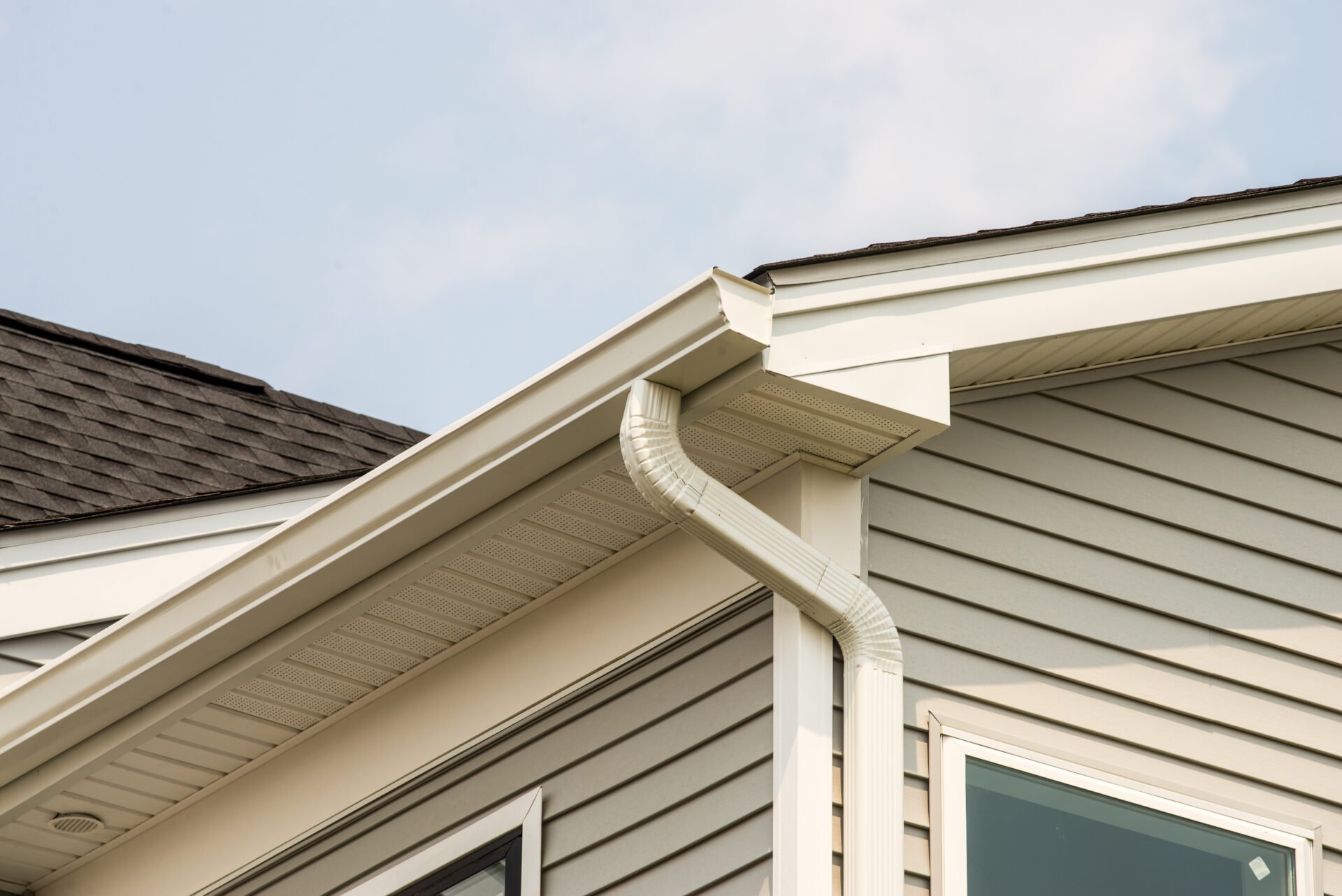Close-up of the corner of a house showing gray siding, white trim, and a white gutter with a downspout against a cloudy sky.