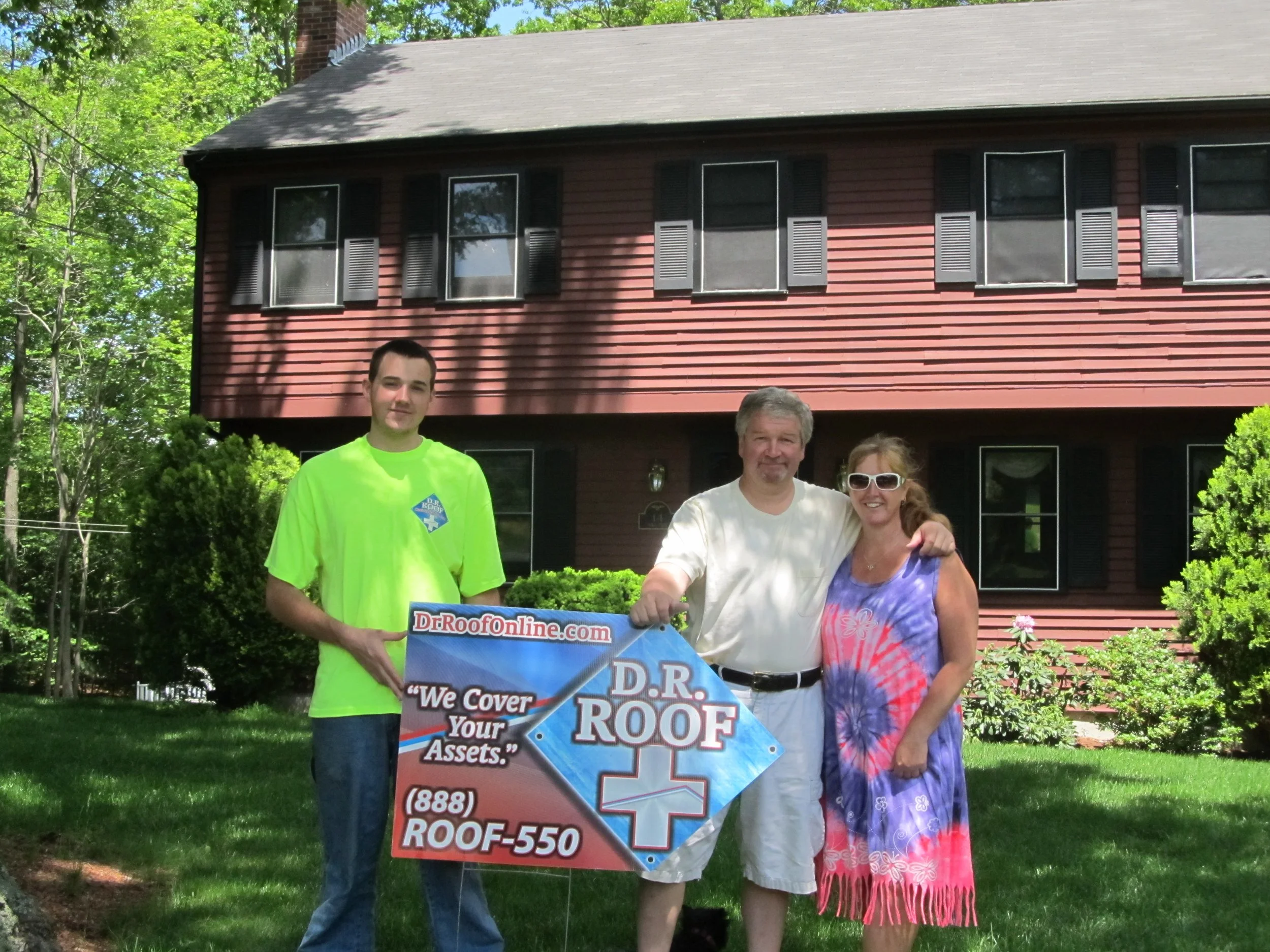 Group of four people standing outdoors in front of a red house with black shutters, holding a sign for a roofing company. Two men and two women, one man in a bright green shirt, another in a white shirt, and one woman wearing sunglasses and a colorful dress.