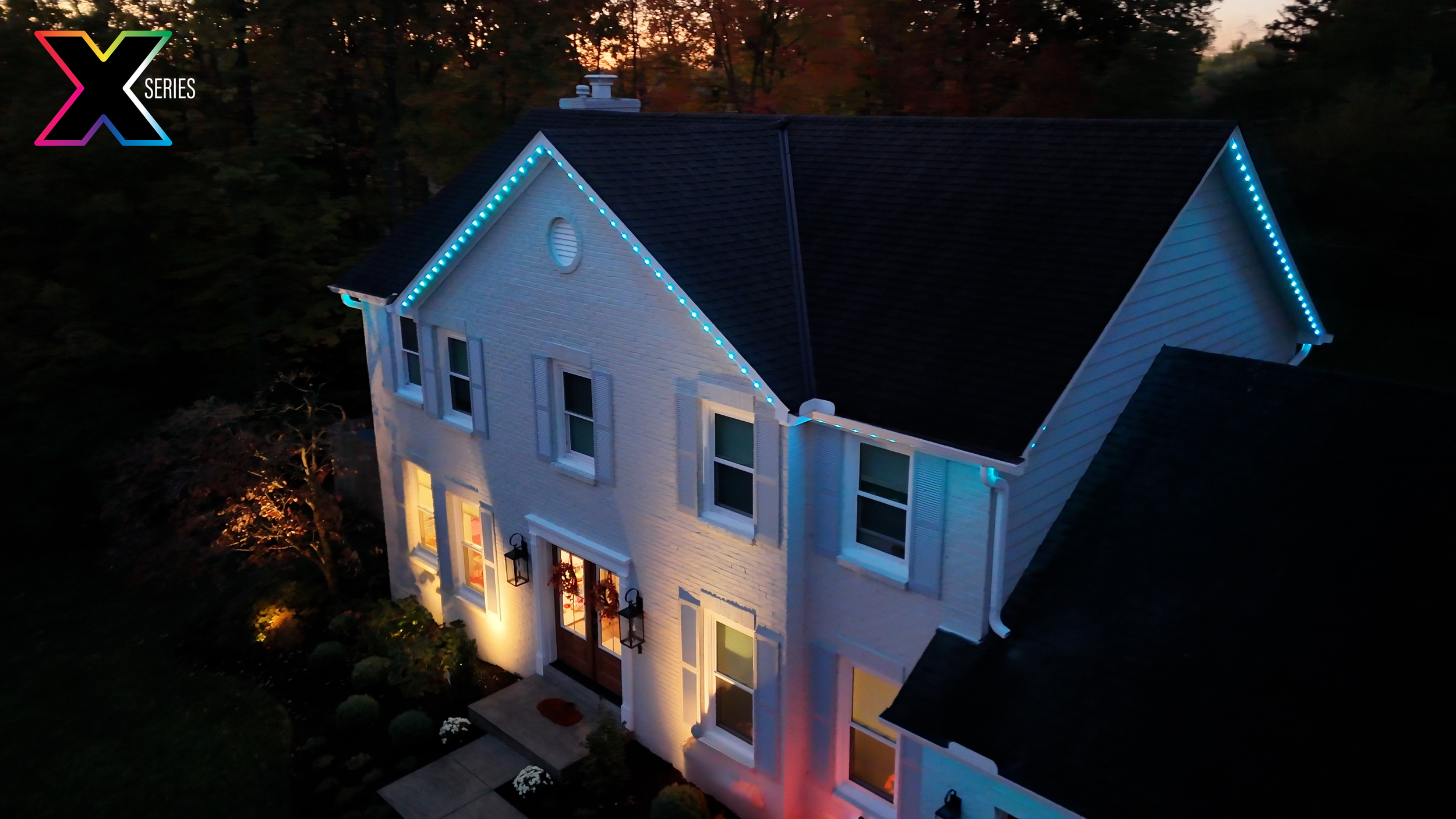 A white two-story house with a porch, illuminated by exterior lights. Blue LED string lights outline the roof's edge. The house is surrounded by trees with fall foliage and illuminated windows. The scene is during dusk or evening.