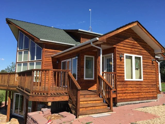 Wooden house with a small front porch, exterior stairs, large windows, and a green roof under a clear blue sky.