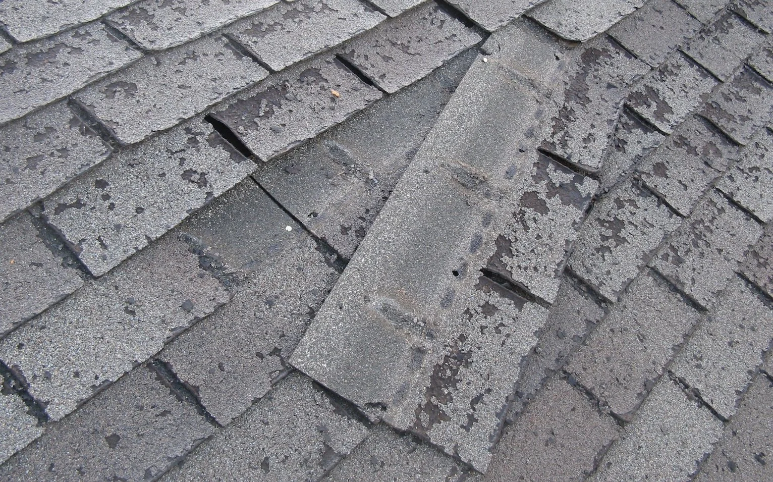 Close-up picture of old, weathered asphalt shingles on a roof, some of which are cracked and broken.
