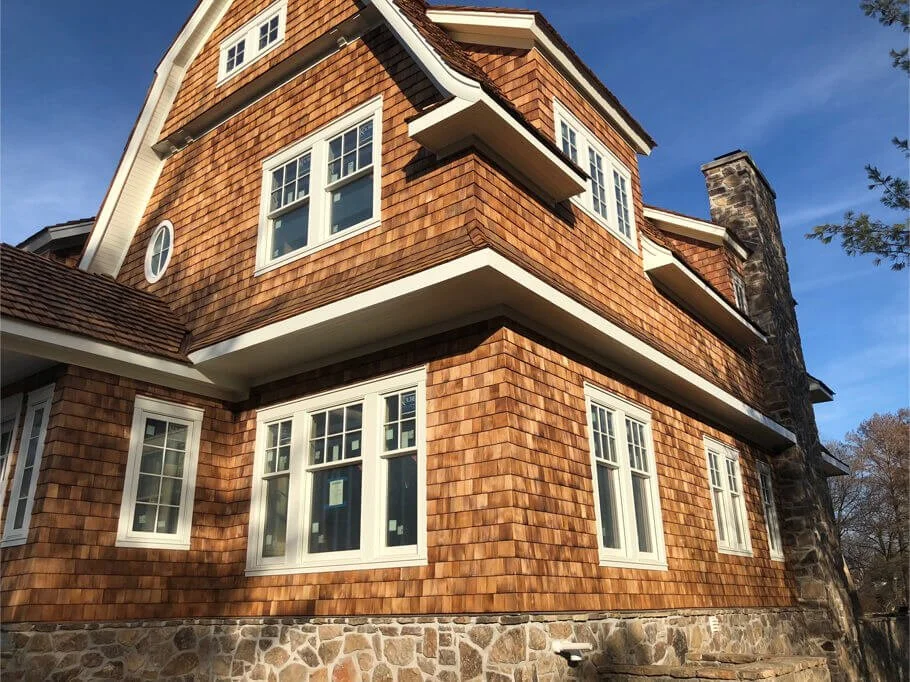 Exterior view of a multi-story house with wooden shingles siding, multiple white-framed windows, a stone foundation, and a chimney on a clear day.