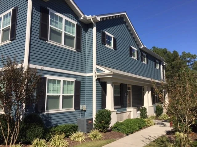 A two-story blue house with white trim and black shutters, with a porch and landscaped bushes and trees in front.