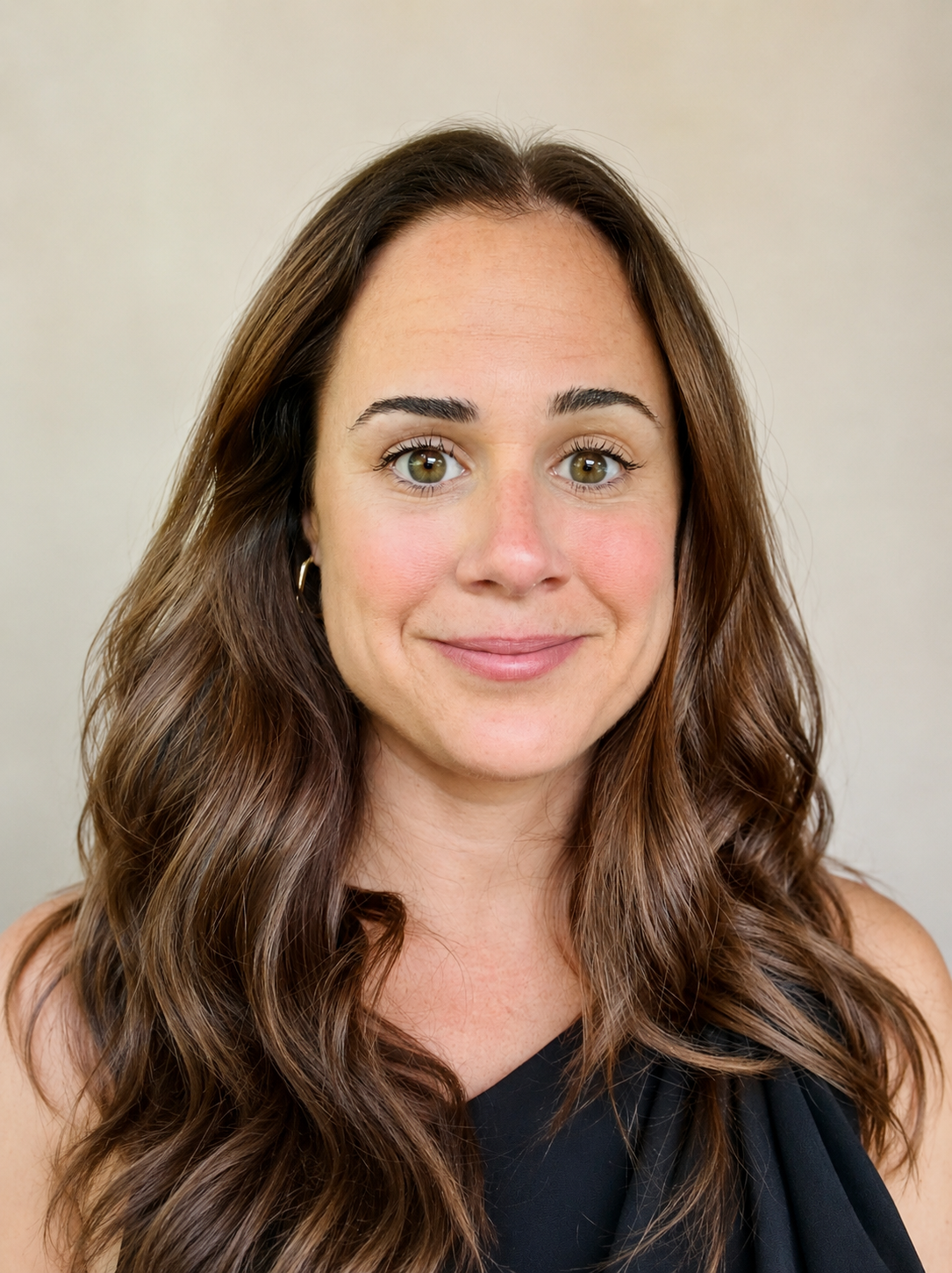 Close-up of a woman with long wavy brown hair, light skin, and green eyes, smiling softly, wearing a black top and hoop earrings.