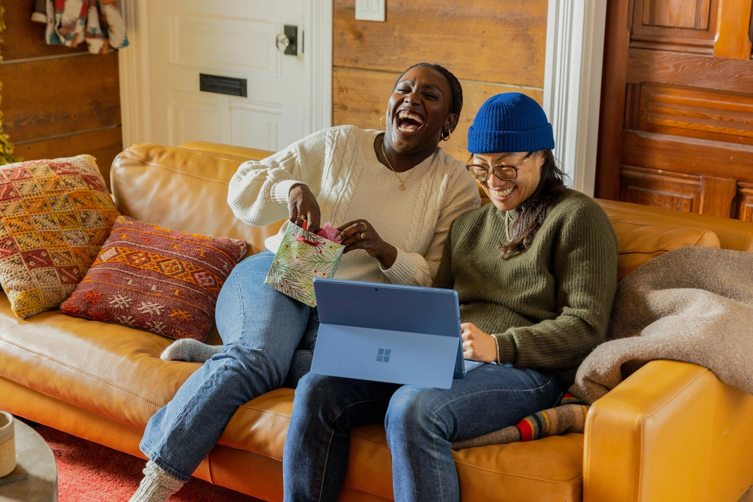 Two women sitting on a tan leather couch, laughing and looking at a laptop. One has short hair and is wearing a white sweater, the other has long hair, glasses, and a blue beanie. There are colorful pillows on the couch and a wooden wall in the background.