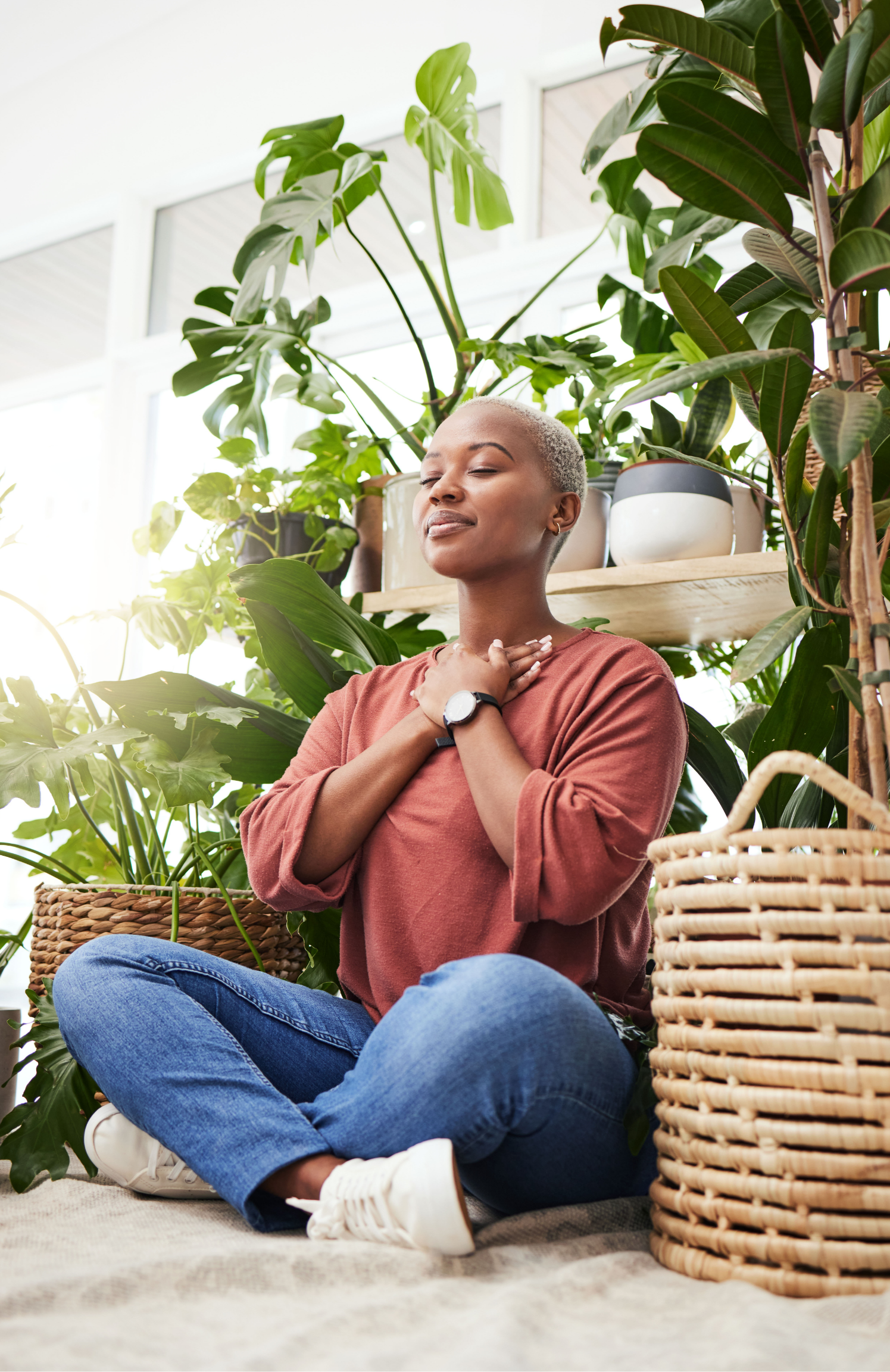 A woman with short platinum blonde hair is sitting cross-legged on the floor with her eyes closed, surrounded by lush green houseplants. She wears a rust-colored top, blue jeans, and white sneakers, and appears to be meditating or relaxing indoors.