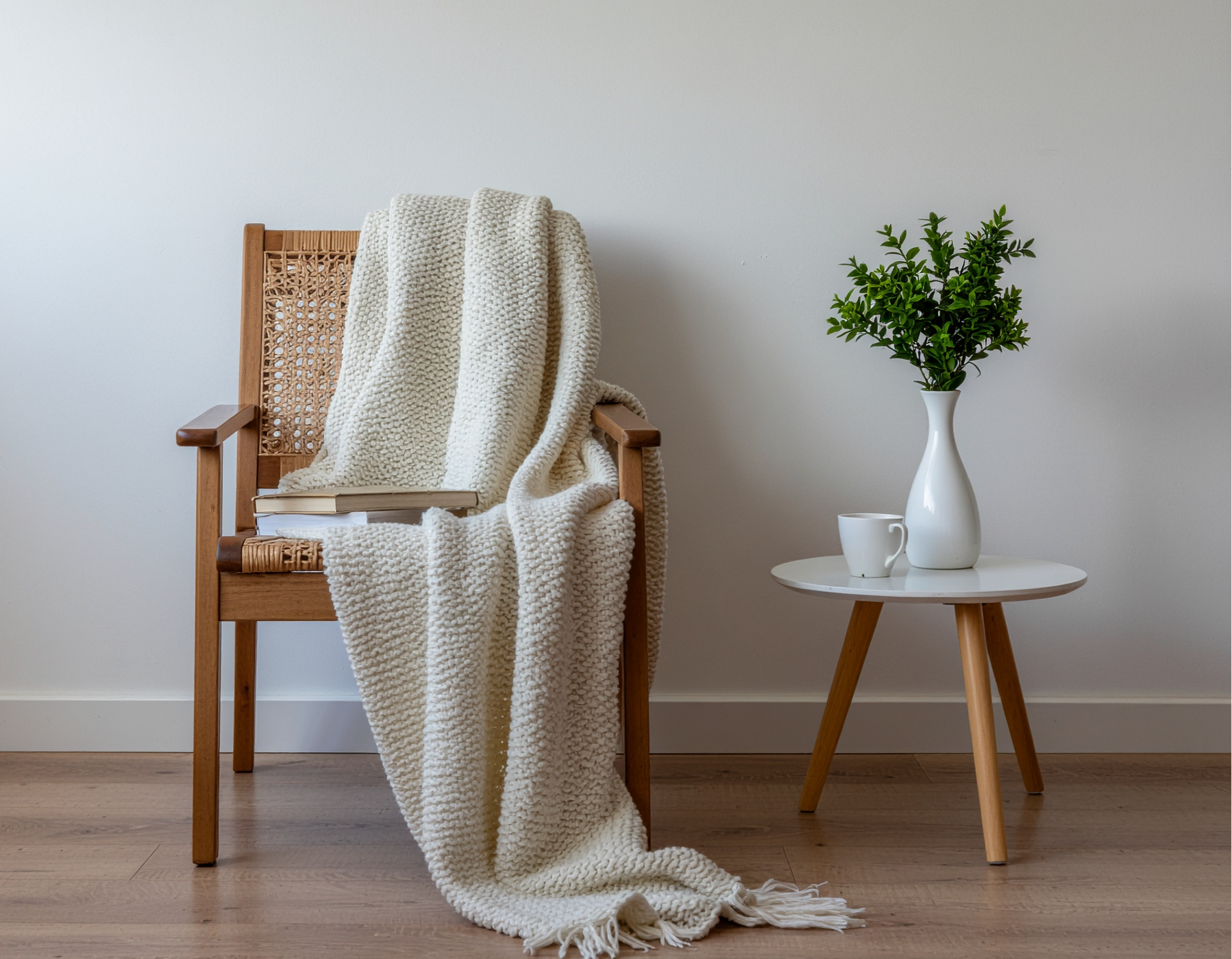 A wooden chair with a woven backrest and armrests, draped with a chunky cream-colored blanket. Next to the chair, on a white round side table, there is a white vase with a green leafy plant and a white mug. The setting is against a plain white wall and wooden floor.
