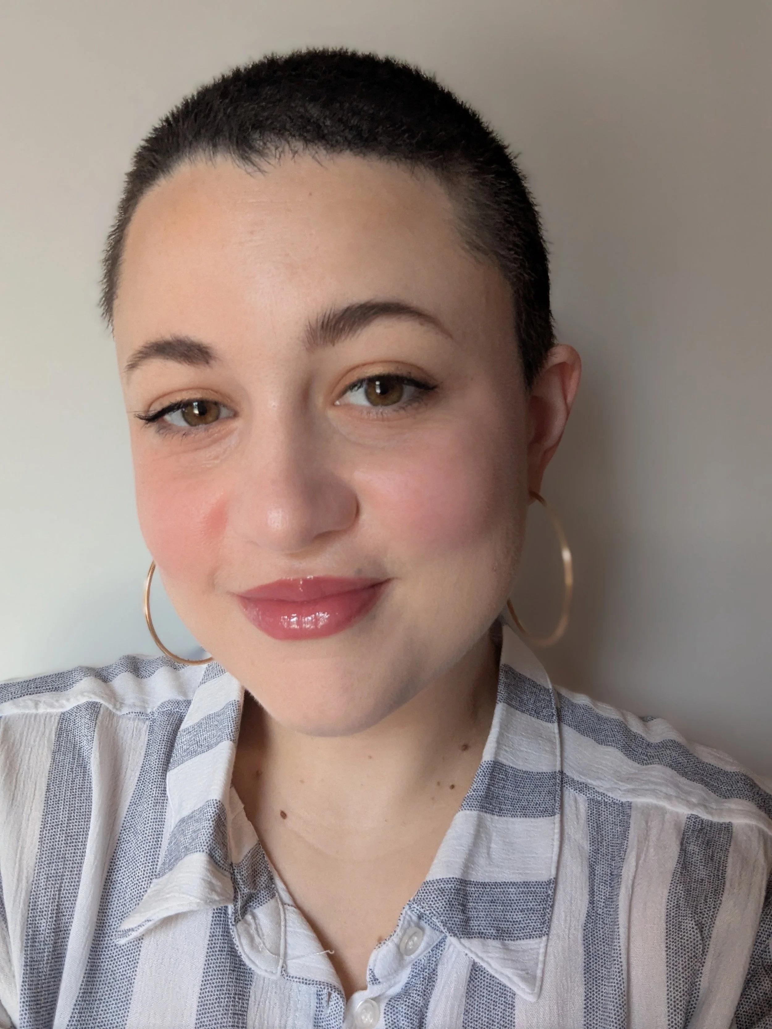 A woman with short dark hair, light skin, and brown eyes, smiling and wearing a striped, button-up shirt and large hoop earrings, standing against a plain wall.