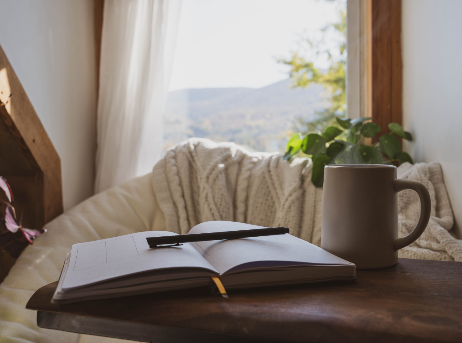 Open notebook with a pen on a wooden table, a ceramic mug, and a cozy bed with a knitted blanket beside a window with white curtains and a view of mountains and trees outside.