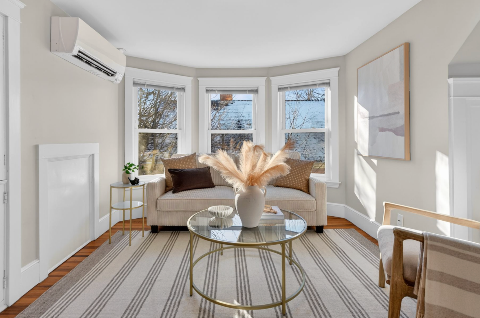 Bright living room with large bay windows, beige sofa with pillows, a gold and glass coffee table with a white vase and pampas grass, striped rug, art on wall, and a small side table with a plant.