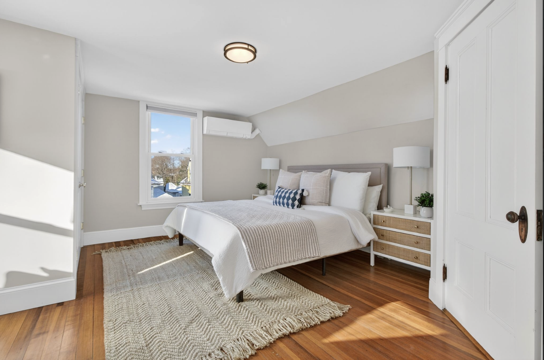 Bright bedroom with white walls, hardwood floor, and a large window. The bed has white bedding, beige pillows, and a decorative pillow. Two nightstands with lamps and small plants are on each side of the bed. A beige rug is on the floor.