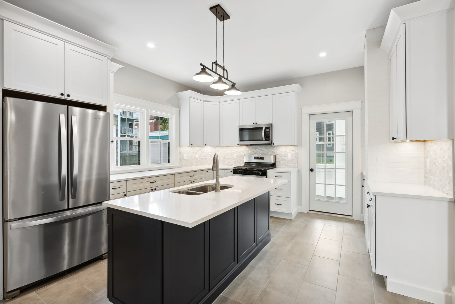 Modern kitchen with white cabinets, a stainless steel refrigerator, black island, built-in microwave, and a door leading outside, brightened by natural and indoor lighting.