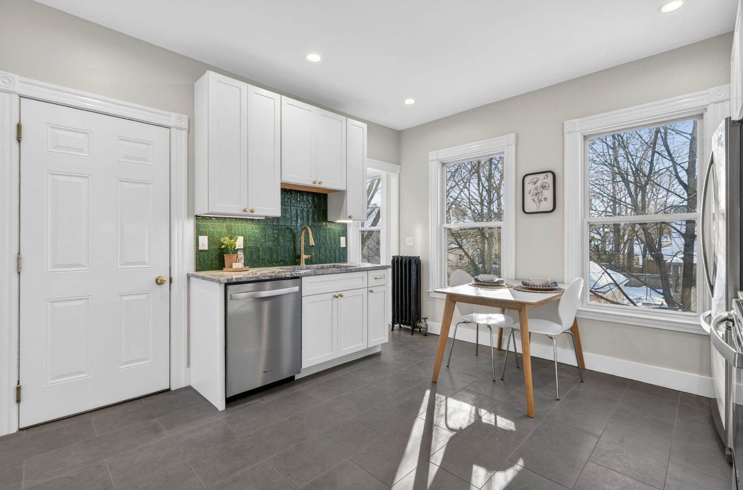Bright kitchen with white cabinets, green tiled backsplash, and a marble countertop. Contains a small plant, sink with gold faucet, and a dining area with a wooden table and white chairs near large windows showing a winter scene outside.