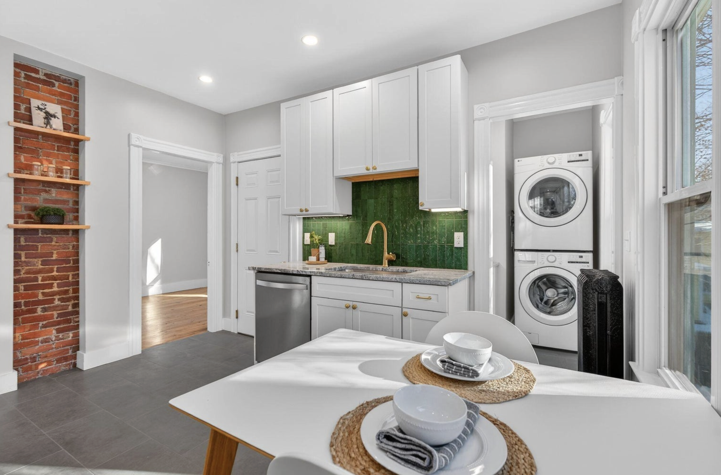 A bright kitchen with white cabinets, green tiled backsplash, a gold faucet, and a stackable washer and dryer next to a window with open blinds. A dining table set with white bowls, napkins, and placemats.