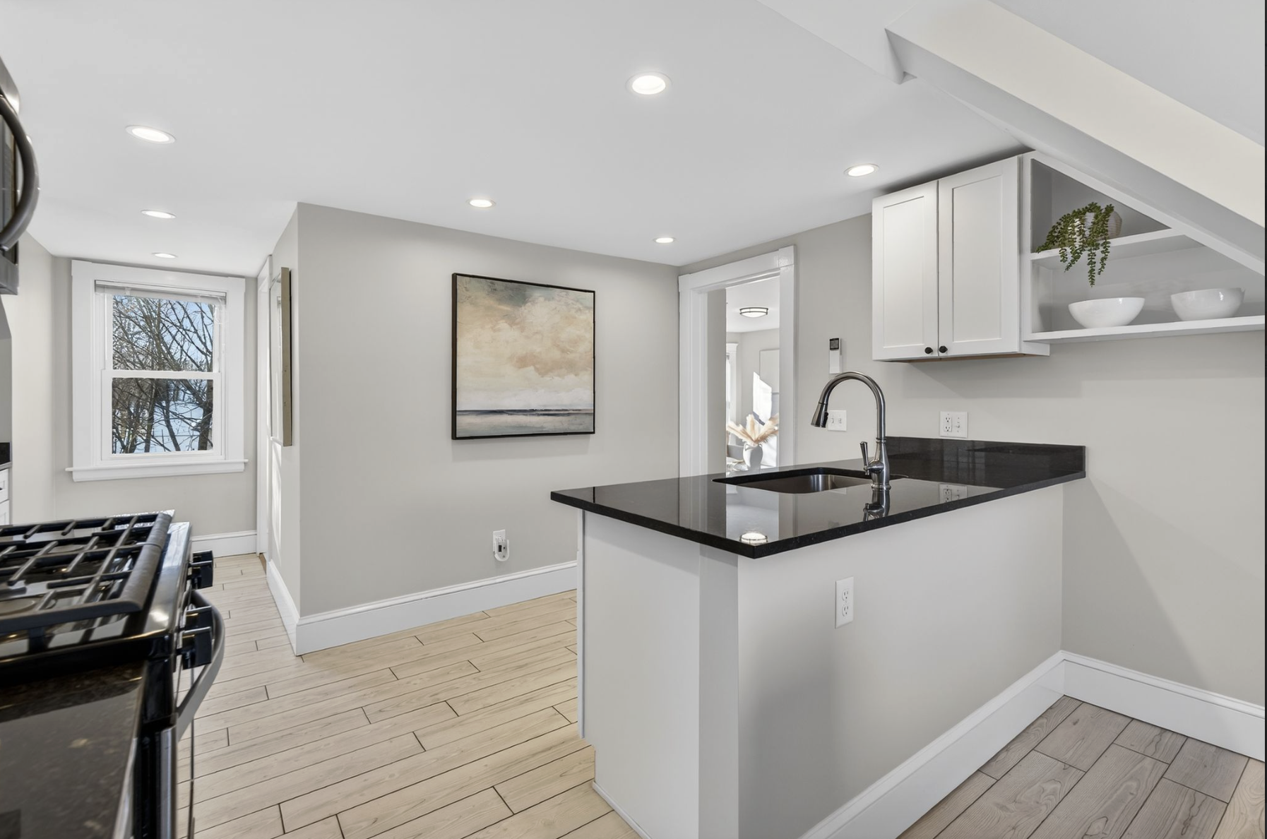 Modern kitchen with white cabinets, black countertop, stainless steel sink and faucet, light hardwood floors, window showing trees, art on the wall, and open space leading to another room.