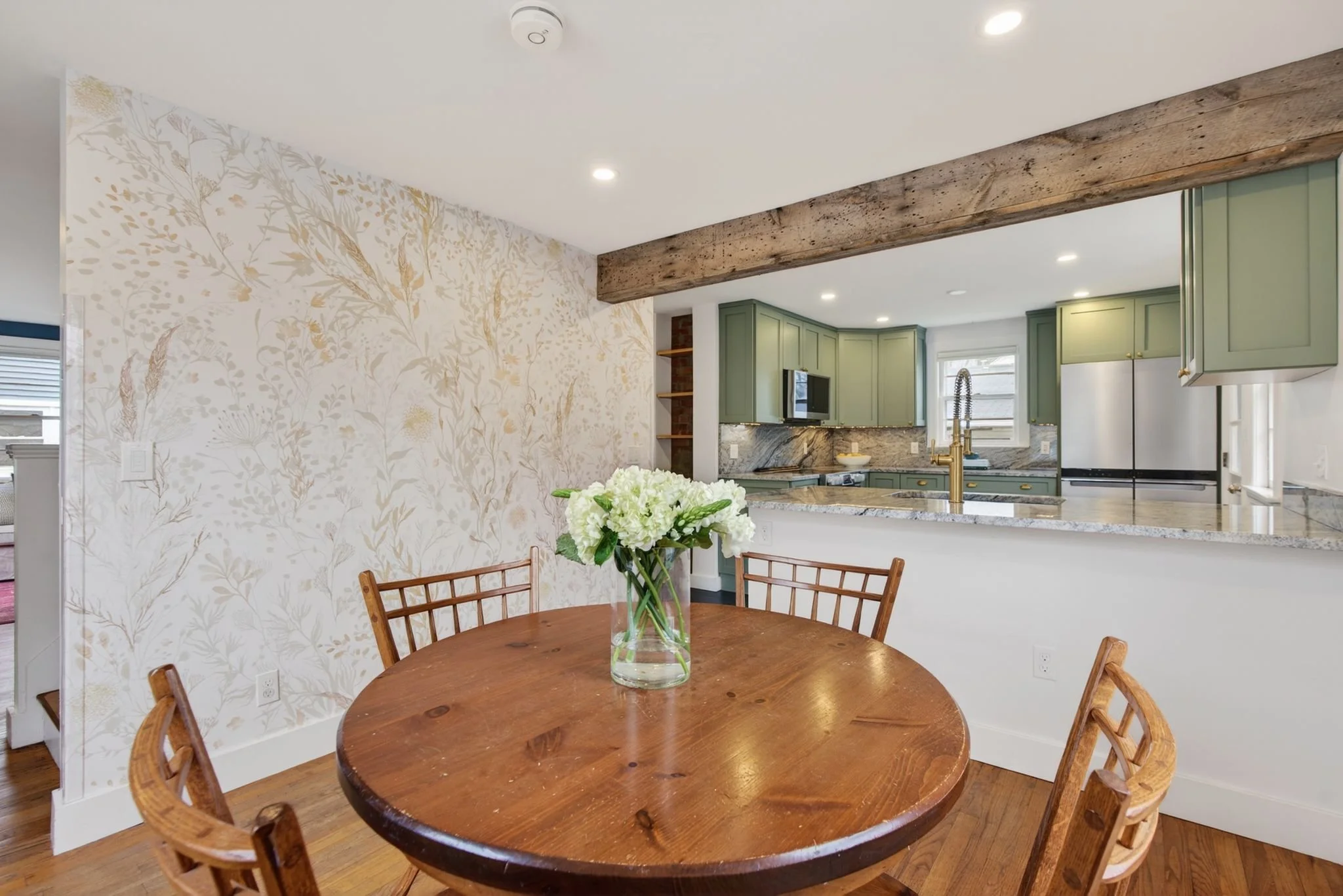 A round wooden dining table with a glass vase holding white flowers, surrounded by wooden chairs, in a dining area with patterned wallpaper, a kitchen with green cabinets, marble countertops, and a wooden beam ceiling.