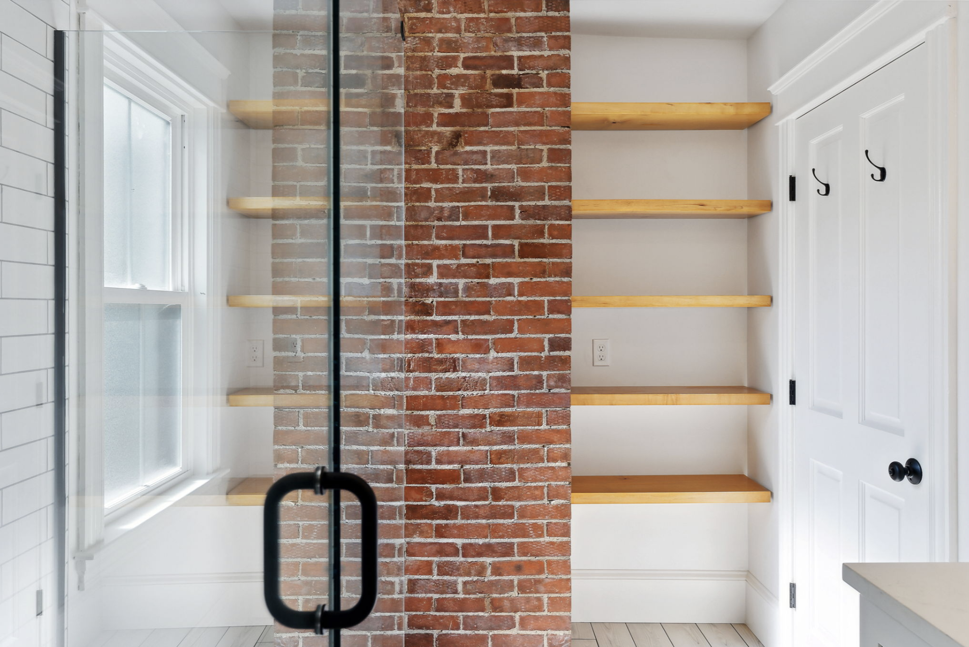Empty pantry with four wooden shelves, a brick wall in the middle, a white wall with hooks, and a window in a bright room.