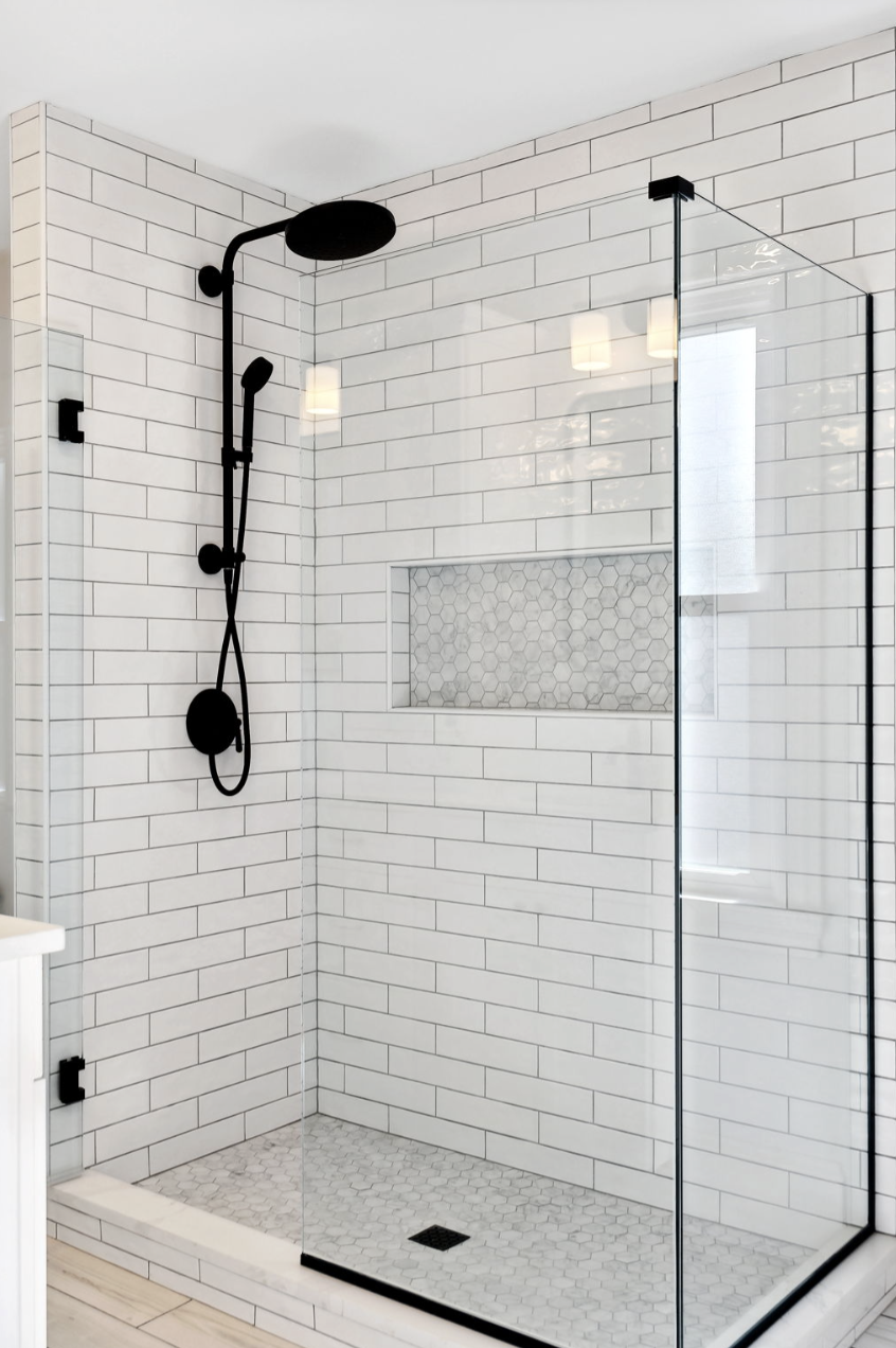 Modern walk-in shower with white subway tile walls, black rainfall and handheld shower fixtures, a built-in niche with hexagon tile, a glass door, and a small hexagon-tile shower floor.