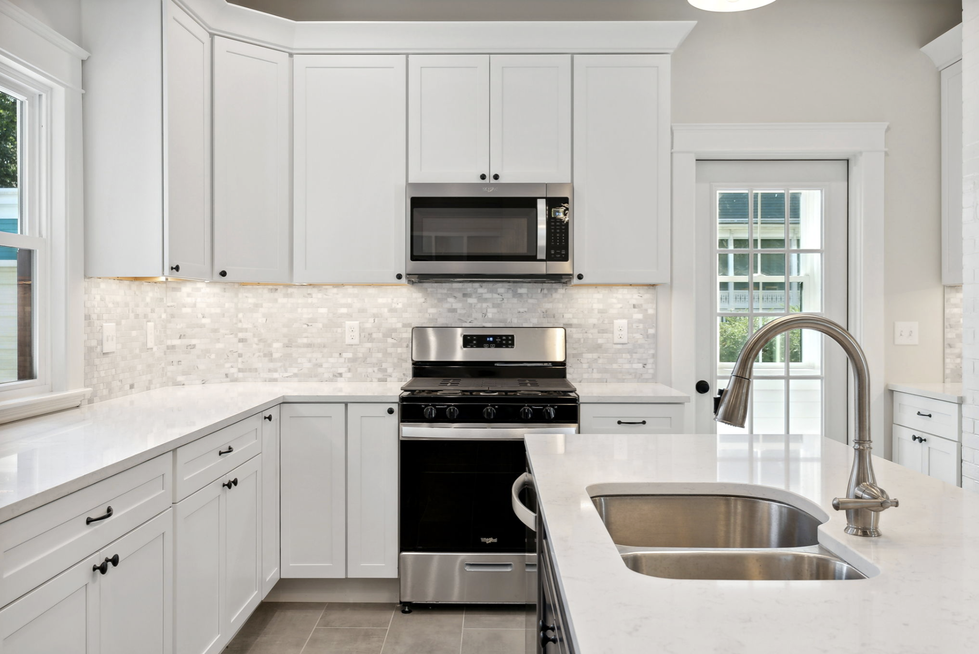 Modern kitchen with white cabinetry, stainless steel microwave and oven, marble countertops, a mosaic tile backsplash, a stainless steel sink with a curved faucet, and a door leading outside with glass panes.