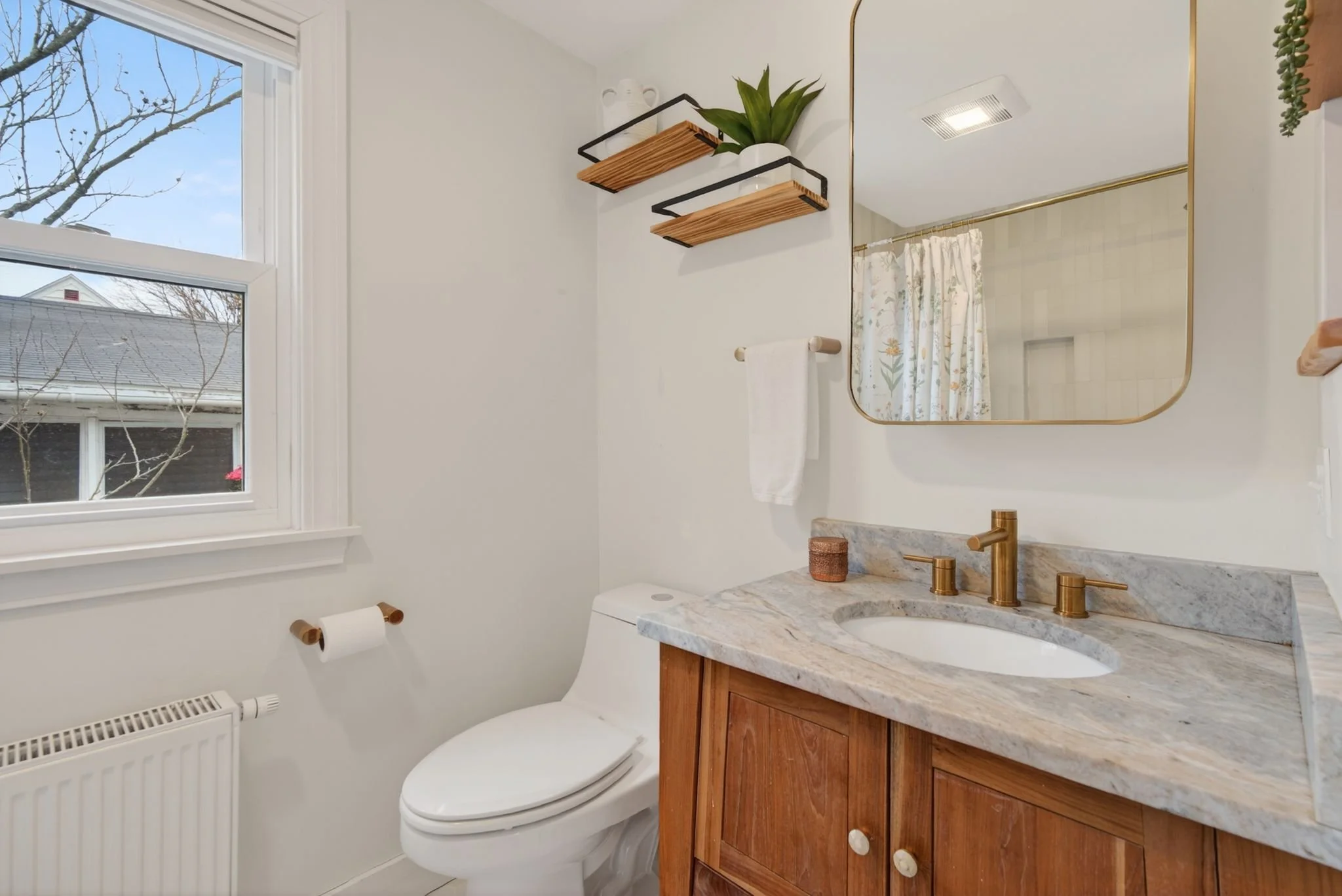 Bathroom with a window, a mirror with a gold frame, a wooden vanity with a marble countertop, a gold-colored faucet, a small potted plant, and a white toilet with a wooden toilet paper holder.