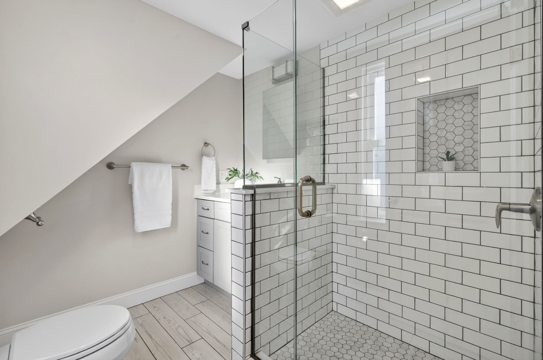 A modern bathroom with a glass shower enclosure, white subway tile walls, and built-in niche with hex tile and a small plant. There are white cabinets, a towel rack with white towels, a potted plant, ceiling vent, and a toilet.