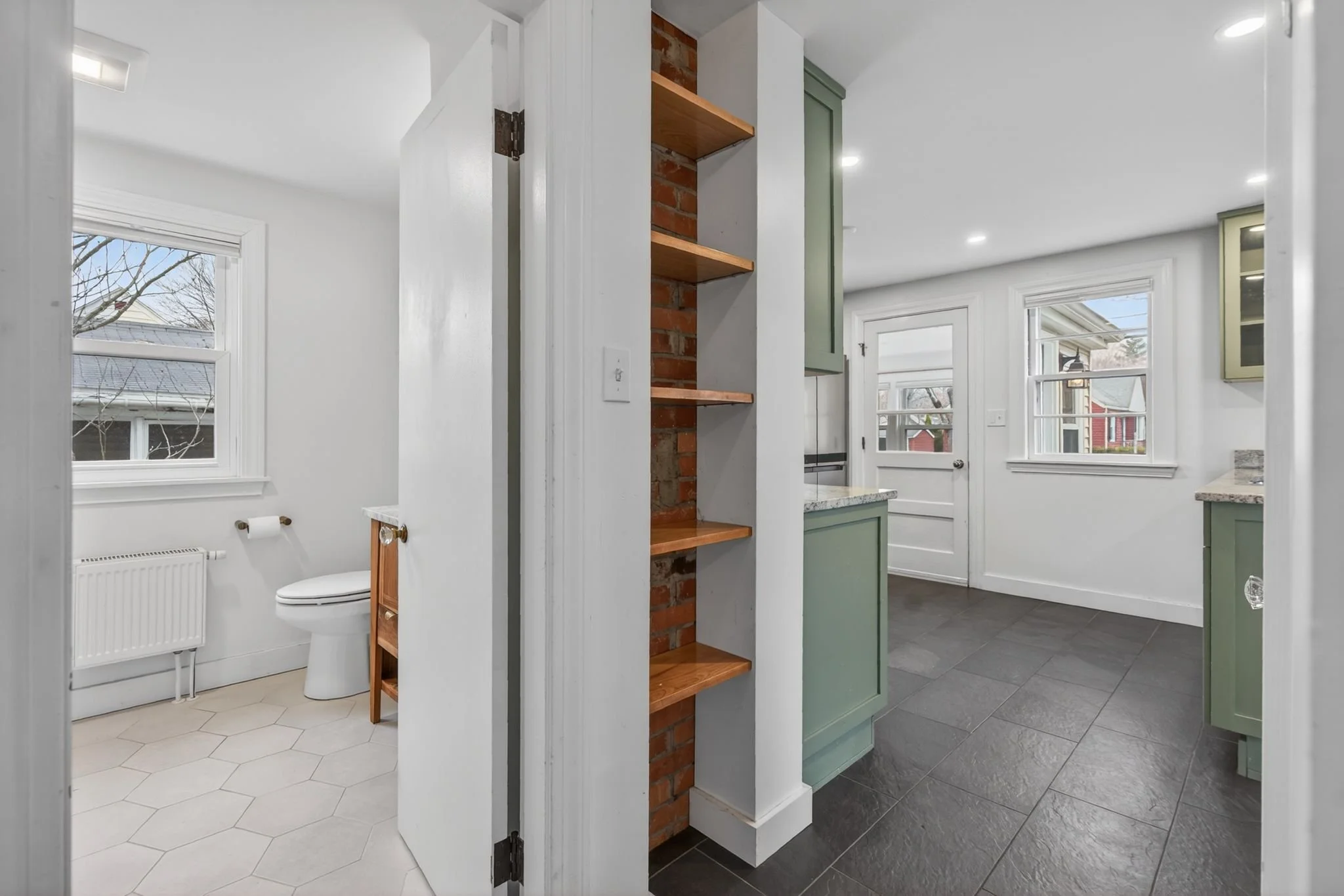 View of a bathroom and kitchen separated by a wall with open shelving. The bathroom has a toilet, a window, and a radiator. The kitchen features green cabinetry, a door, and large windows, with a mix of light and dark flooring.